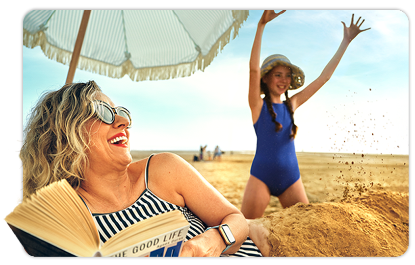 Image of a woman reading on the beach with her granddaughter playing in the sand 